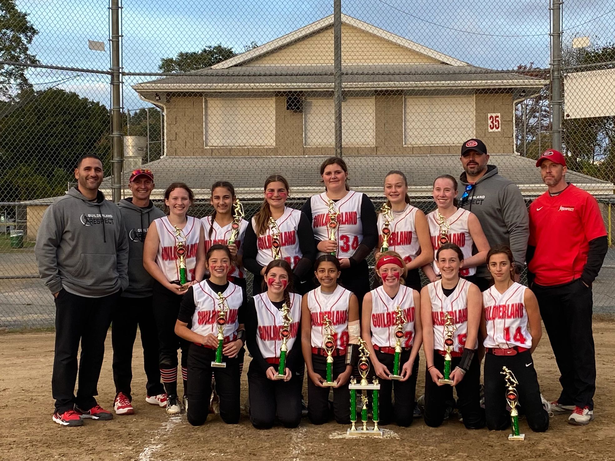 Girls softball team posing with trophies on a baseball field.