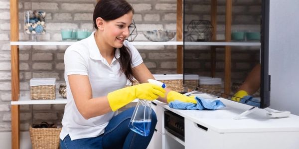 Woman wearing yellow gloves cleaning a TV stand with a spray bottle and cloth.