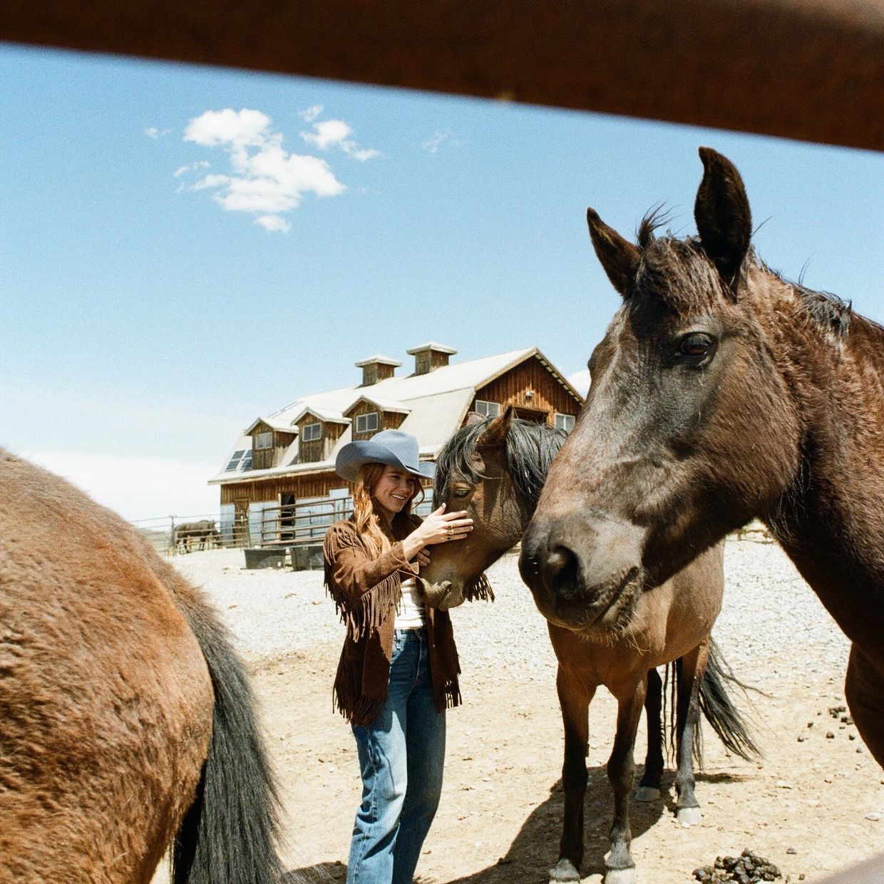 A woman in a cowboy hat petting a horse on a sunny day at a ranch.