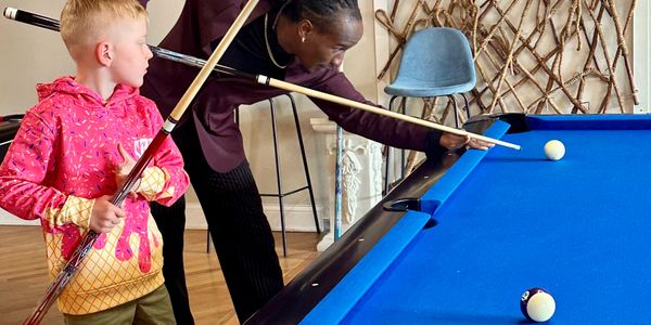 An adult teaches a child to play pool indoors with a blue pool table.