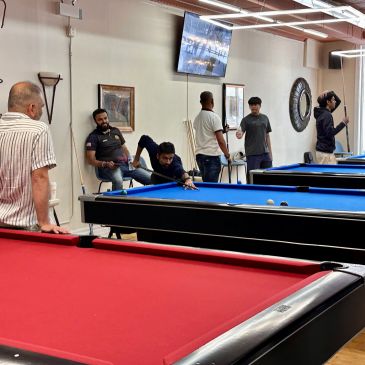 Group of men playing and watching pool in a billiards room with red and blue tables.