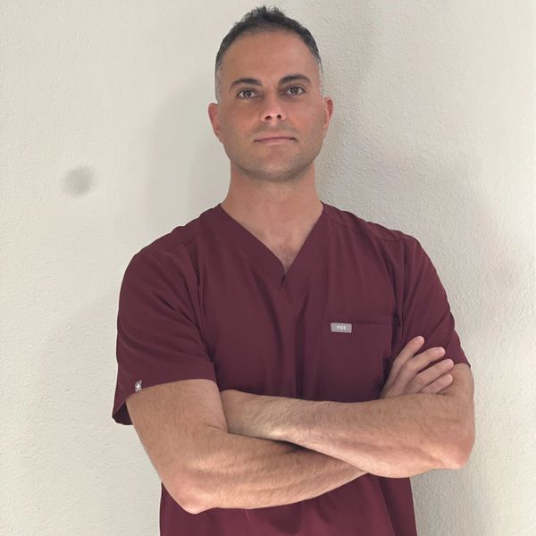 Man in maroon medical scrubs with arms crossed, standing against a plain wall.