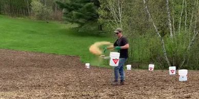 Man spreading granular fertilizer on a field using a handheld spreader.