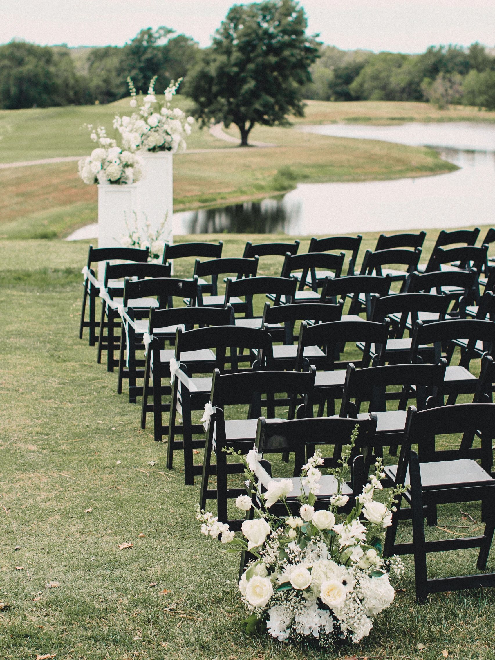Multiple flower displays and chairs set up in rows, outside over looking a lake, for a wedding. 