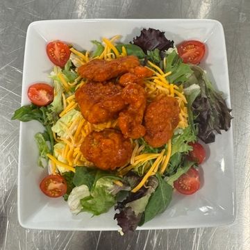 A white square bowl sitting on a metal table filled with a leafy green buffalo chicken salad.