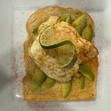 A white plate topped with a large piece of toast covered in sliced avocado, an egg, lime and paprika