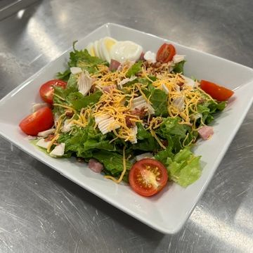 a white square  salad bowl filled with green lettuce, tomato, egg, diced ham, and shredded cheese.