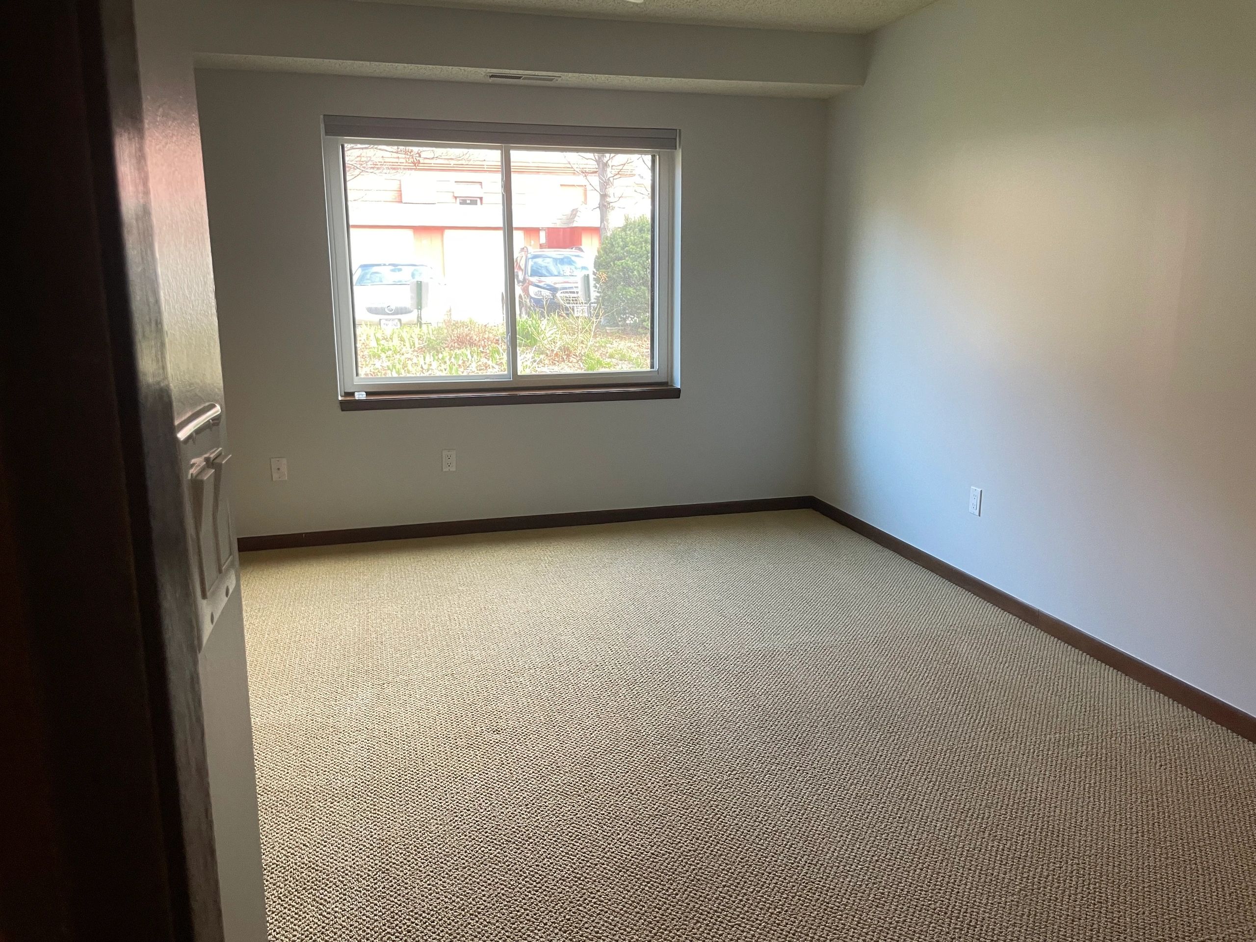 Empty room with beige carpet and a window showing parked cars outside.