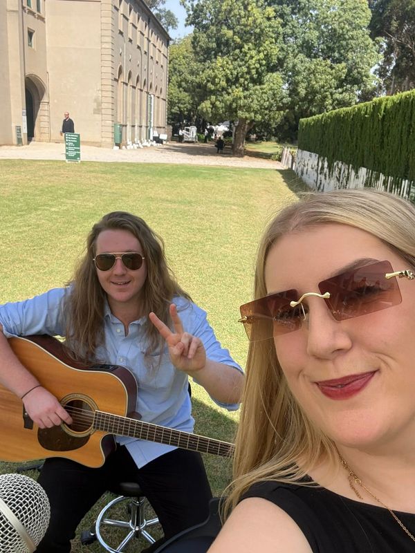 Two people outdoors, one playing guitar and making a peace sign, the other taking a selfie with sunglasses.