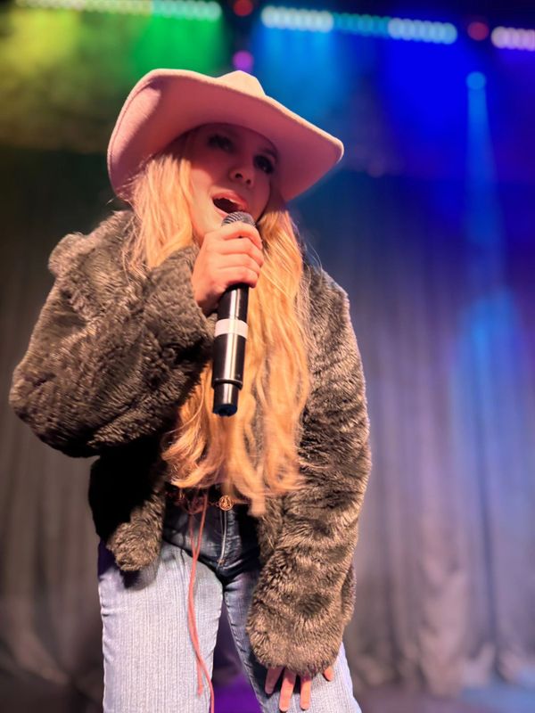 Singer in a pink cowboy hat and fur coat performing on stage.