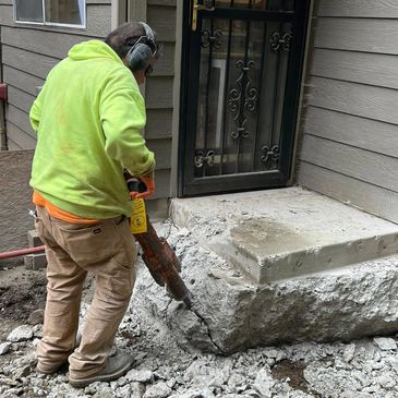 Worker using jackhammer to break concrete near a house entrance.