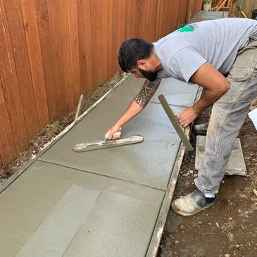 A man smooths wet concrete on a sidewalk beside a wooden fence.