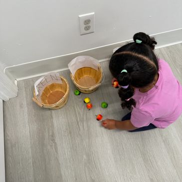 A young girl sorting colorful toy fruits into labeled baskets on the floor.