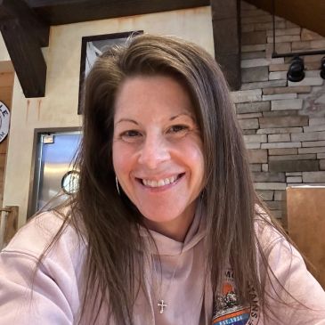 Smiling woman with long hair wearing a light pink hoodie indoors.
