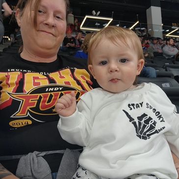 A woman and baby at a sports event, baby wearing a 'Stay Spooky' shirt.