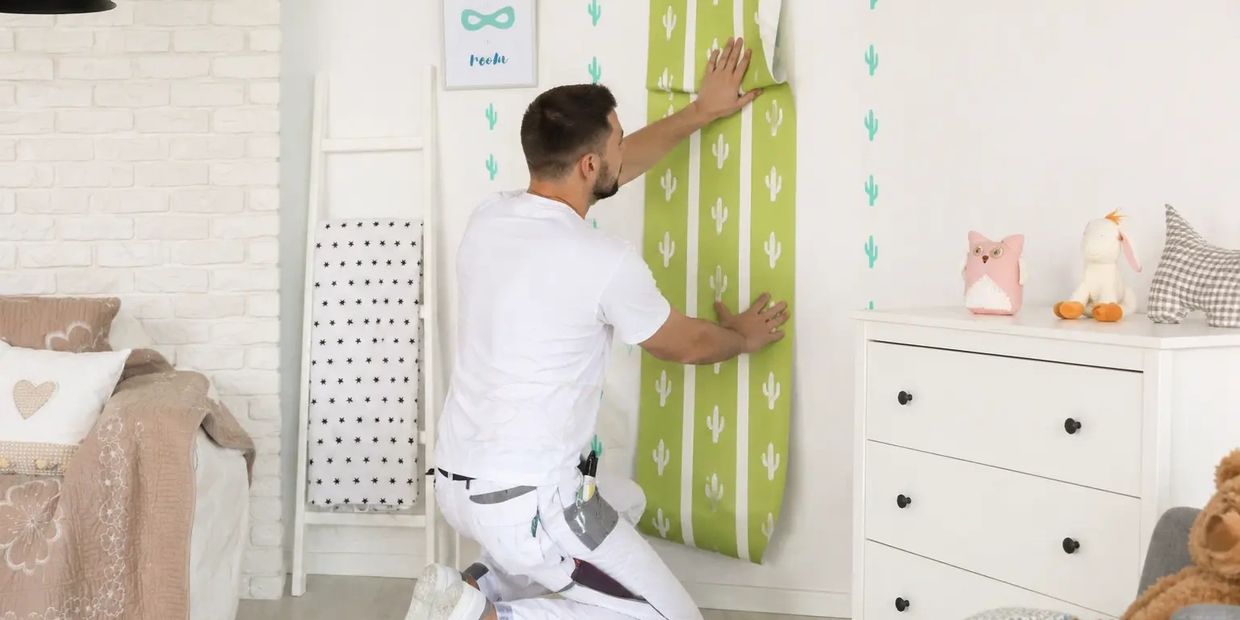 Man applying green cactus-patterned wallpaper in a cozy, white nursery room.