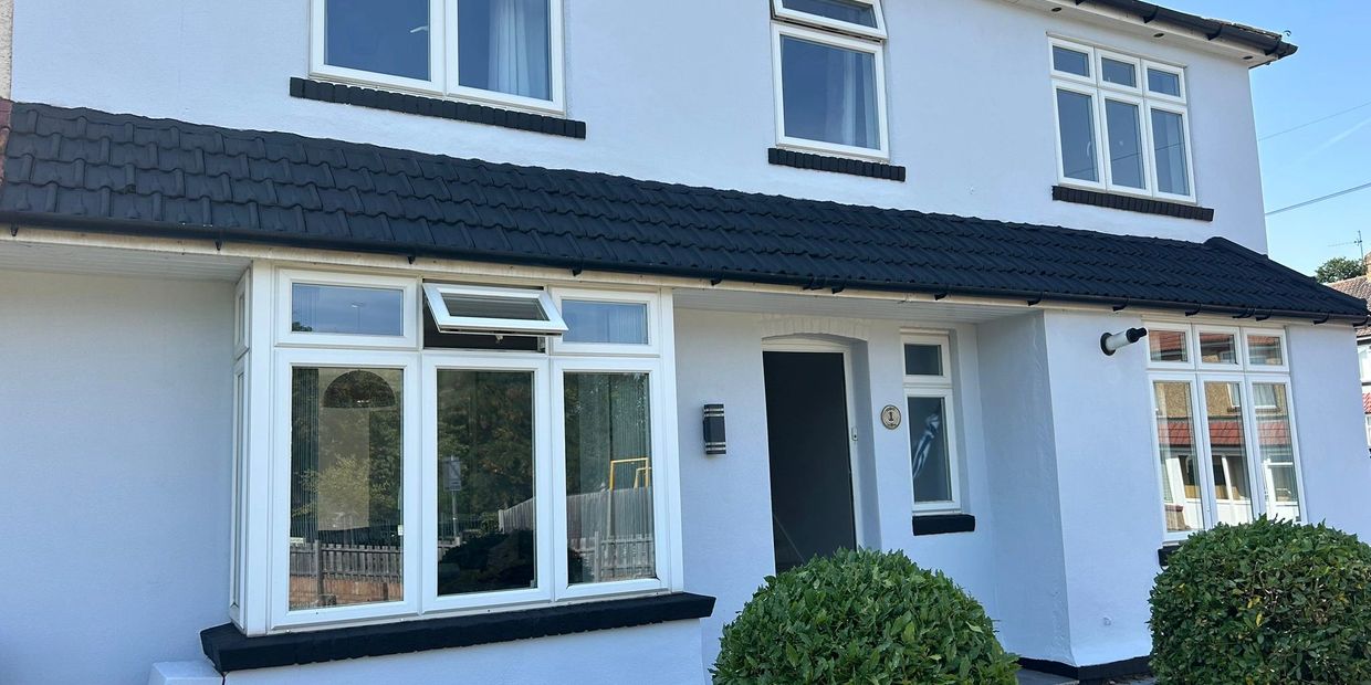White two-story house with black roof tiles and green bushes in front.