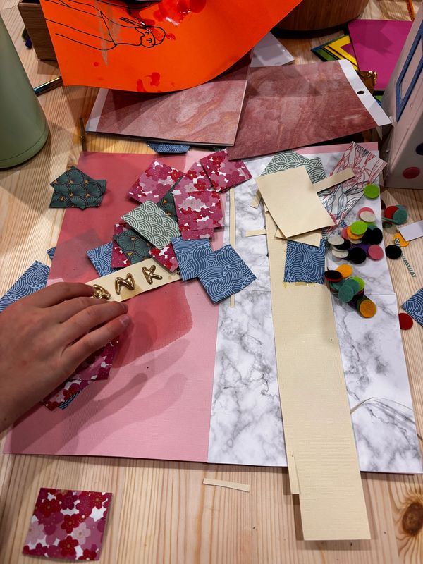 Hand arranging patterned paper pieces on a crafting table with various materials scattered.