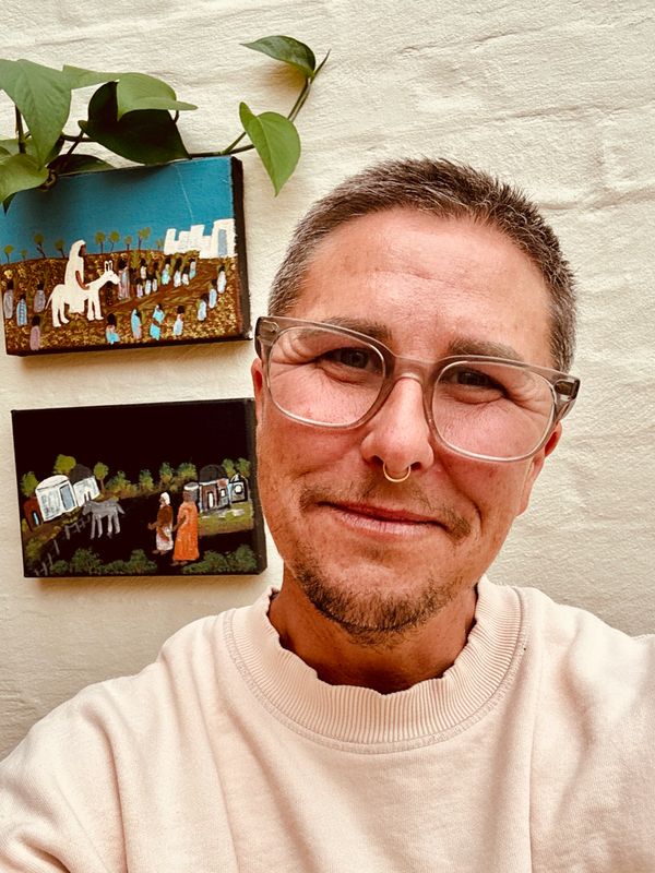 Smiling person with glasses and septum piercing poses in front of wall art.