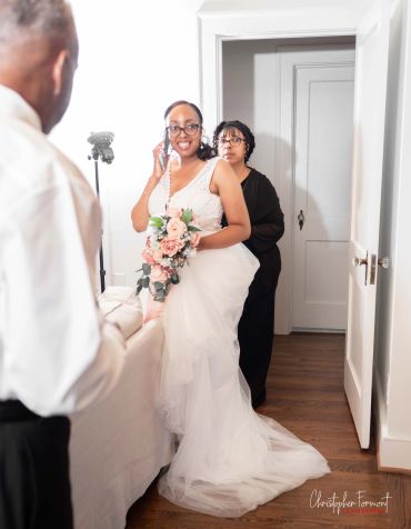 Bride in white dress holding flowers, talking on phone, with two people nearby.