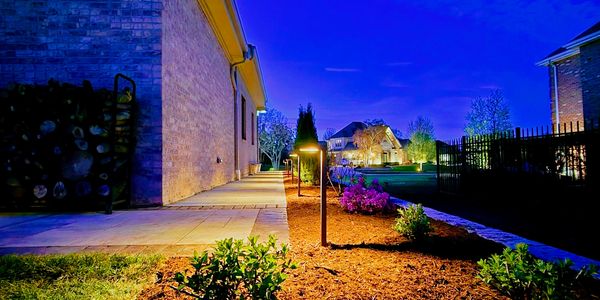 Outdoor pathway lighting illuminating plants beside a brick house at dusk.