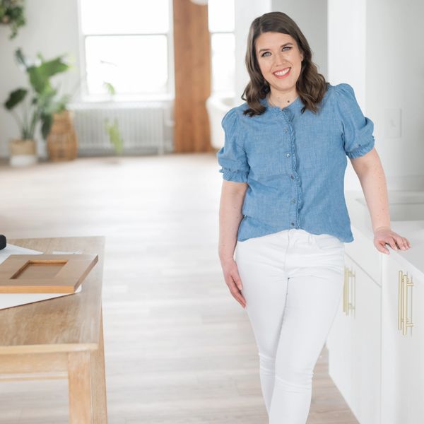 Smiling woman in blue blouse and white pants leaning on kitchen counter.