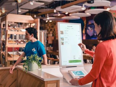 Woman using a self-service kiosk at a store with a staff member in the background.