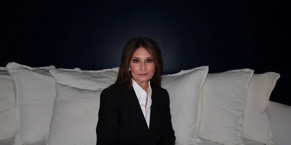 Woman in a black blazer sitting on a white couch against a dark background.