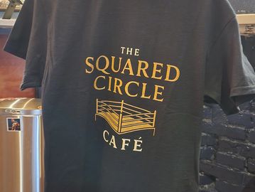 Black t-shirt with gold text and a wrestling ring graphic.