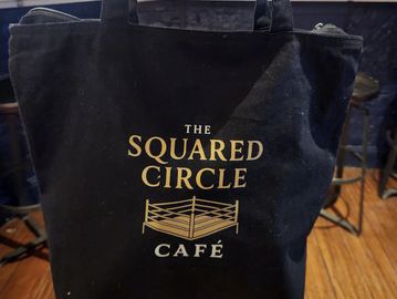 Black bag with 'The Squared Circle Café' logo featuring a wrestling ring.