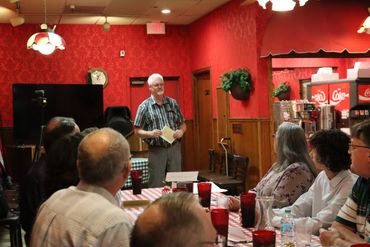 Man speaking to a group in a cozy red-walled restaurant.