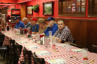 Men sitting at a long table with red checkered tablecloth in a diner.