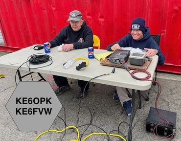 Two men operate amateur radio equipment outdoors at a table with call signs displayed.