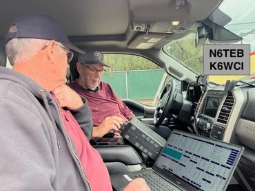 Two men using ham radio and laptop inside a vehicle.