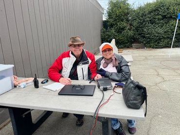 Two elderly people sitting at an outdoor table with electronics and papers.