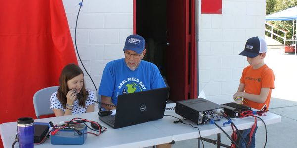 An elderly man and two children operate amateur radio equipment outdoors.