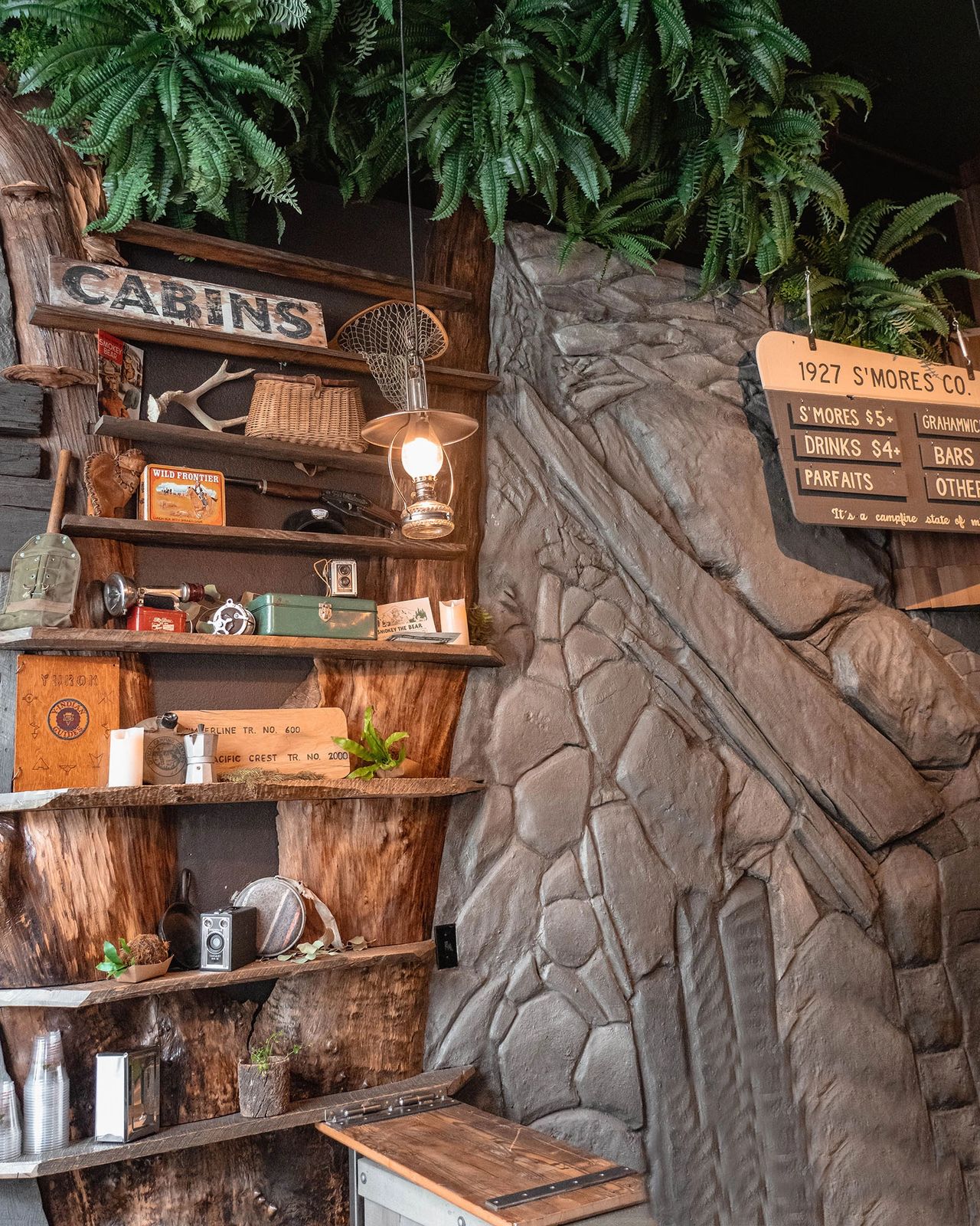 The cozy interior of 1927 S’mores Company in Portland, Oregon, designed to evoke nostalgic summer camp vibes. Wooden shelves decorated with vintage camping gear, lanterns, antlers, and old cameras create a rustic, cabin-like atmosphere. Ferns hang from above, and a menu board lists s’mores and drinks, adding to the playful, outdoorsy theme. This unique dessert café is a must-visit for travelers looking for a warm and whimsical experience in downtown Portland. Photo by Nastasia Yakoub