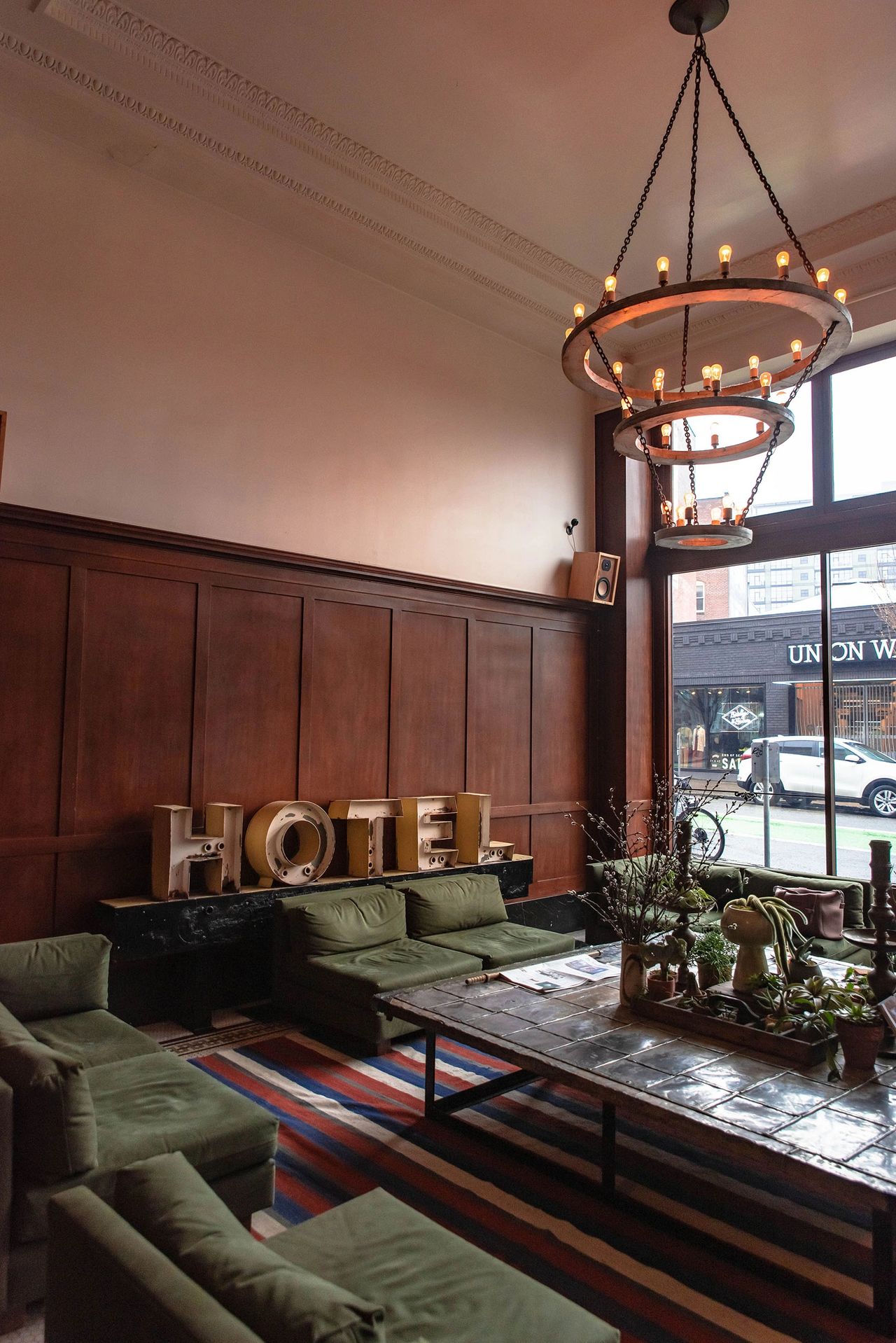 The stylish lobby of the Ace Hotel in Portland, Oregon, featuring olive-green mid-century sofas, a large tiled coffee table, vintage marquee “HOTEL” letters, and warm wood-paneled walls. A modern chandelier with exposed bulbs hangs from the ceiling, while large windows let in natural light from the street outside. The Ace Hotel blends hip, vintage-inspired design with a laid-back atmosphere, making it a popular choice for creatives and travelers exploring Portland.