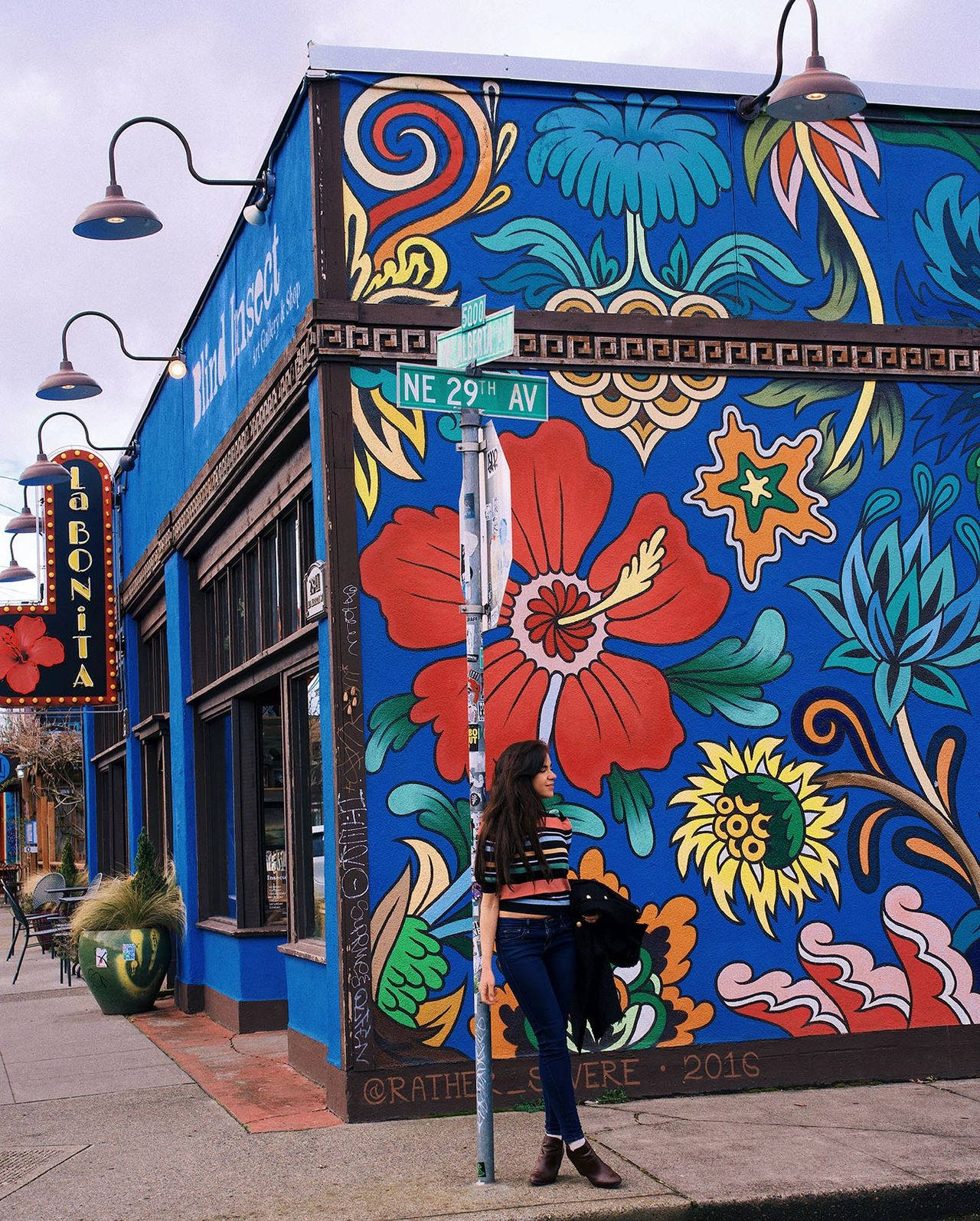 A woman stands on the corner of NE 29th Avenue and Alberta Street in Portland, Oregon, in front of a vibrant floral mural painted on a bright blue building. The bold street art and colorful details reflect the creative spirit of the Alberta Arts District, one of Portland’s most eclectic neighborhoods. Known for its murals, trendy cafés, and local boutiques, this area is a top destination for visitors seeking street art and cultural experiences. Photo by Nastasia Yakoub