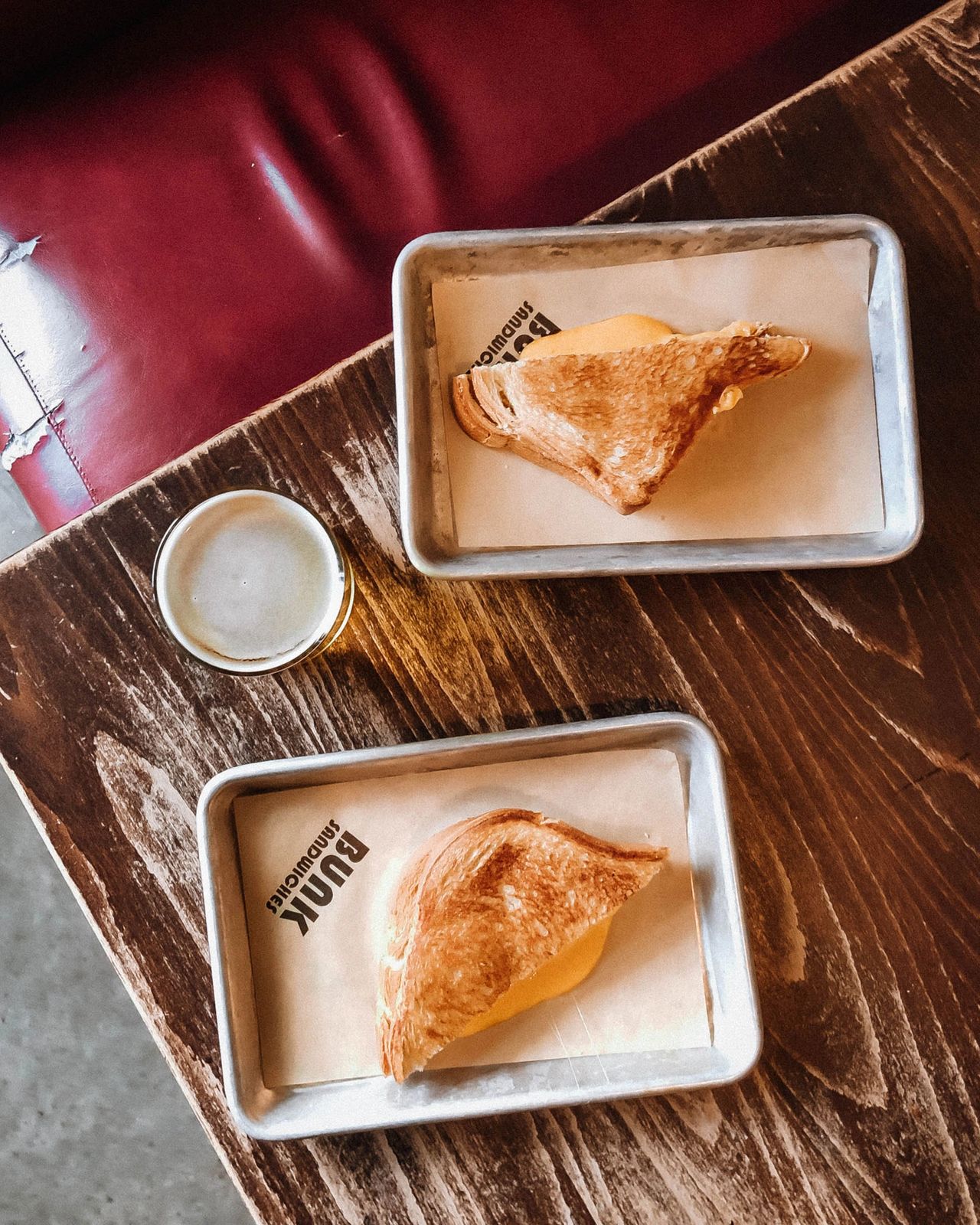 Overhead view of two golden, melty grilled cheese sandwiches and a cold beer on a rustic wooden table at Bunk Sandwiches in Portland, Oregon. Known for its perfectly toasted sandwiches, relaxed atmosphere, and local charm, this Portland favorite is a must-visit for comfort food lovers exploring the city’s thriving food scene. Photo by Nastasia Yakoub
