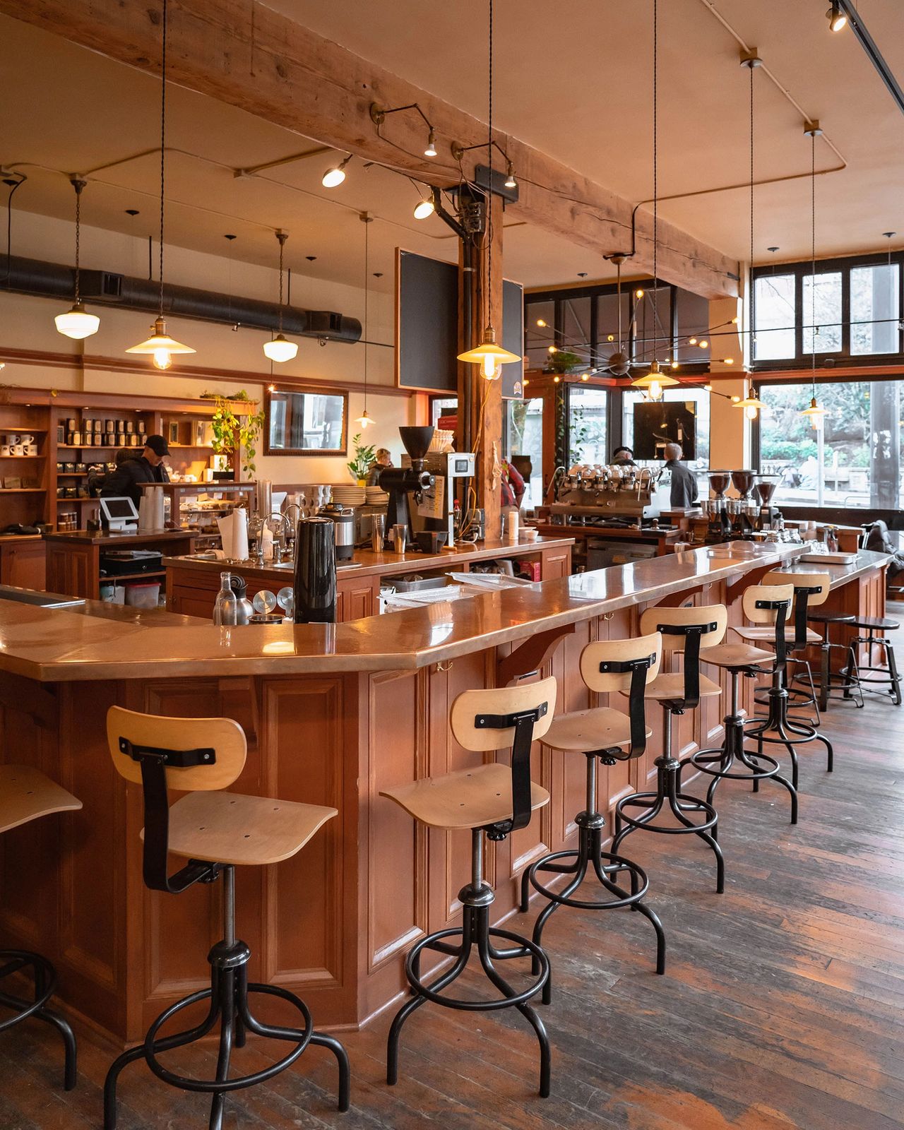 The cozy interior of Case Study Coffee in downtown Portland, Oregon, featuring a long wooden bar lined with industrial-style stools, warm pendant lighting, and exposed wood beams. The café has a welcoming, vintage-meets-modern atmosphere with baristas preparing coffee behind the counter and natural light streaming through large windows. Case Study Coffee is a beloved Portland spot known for its quality espresso, beautiful design, and relaxed vibe. Photo by Nastasia Yakoub