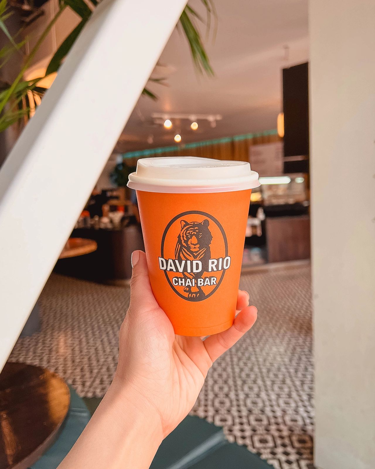 Hand holding a vibrant orange David Rio Chai Bar cup inside a stylish San Francisco café.