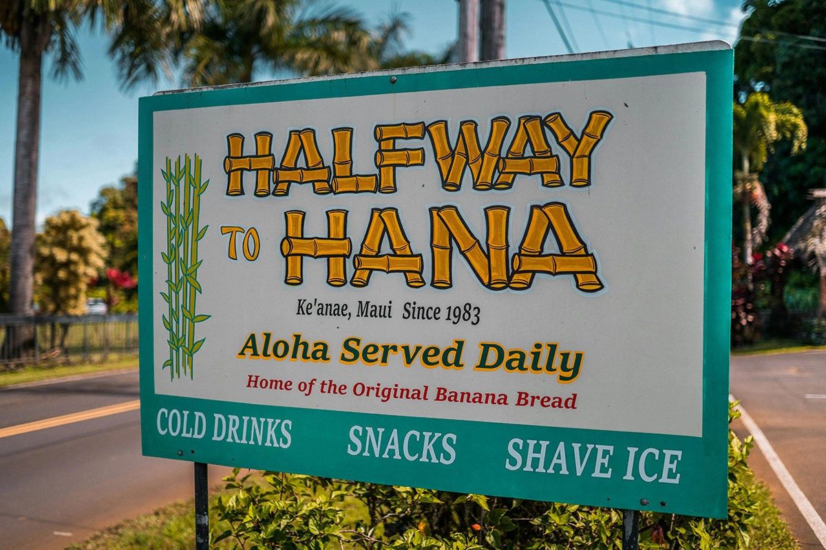Iconic Halfway to Hana roadside stand sign in Ke'anae, Maui. Text reads: Halfway to Hana, Ke'anae Maui Since 1983, Aloha Served Daily, Home of the Original Banana Bread, Cold Drinks, Snacks, Shave Ice