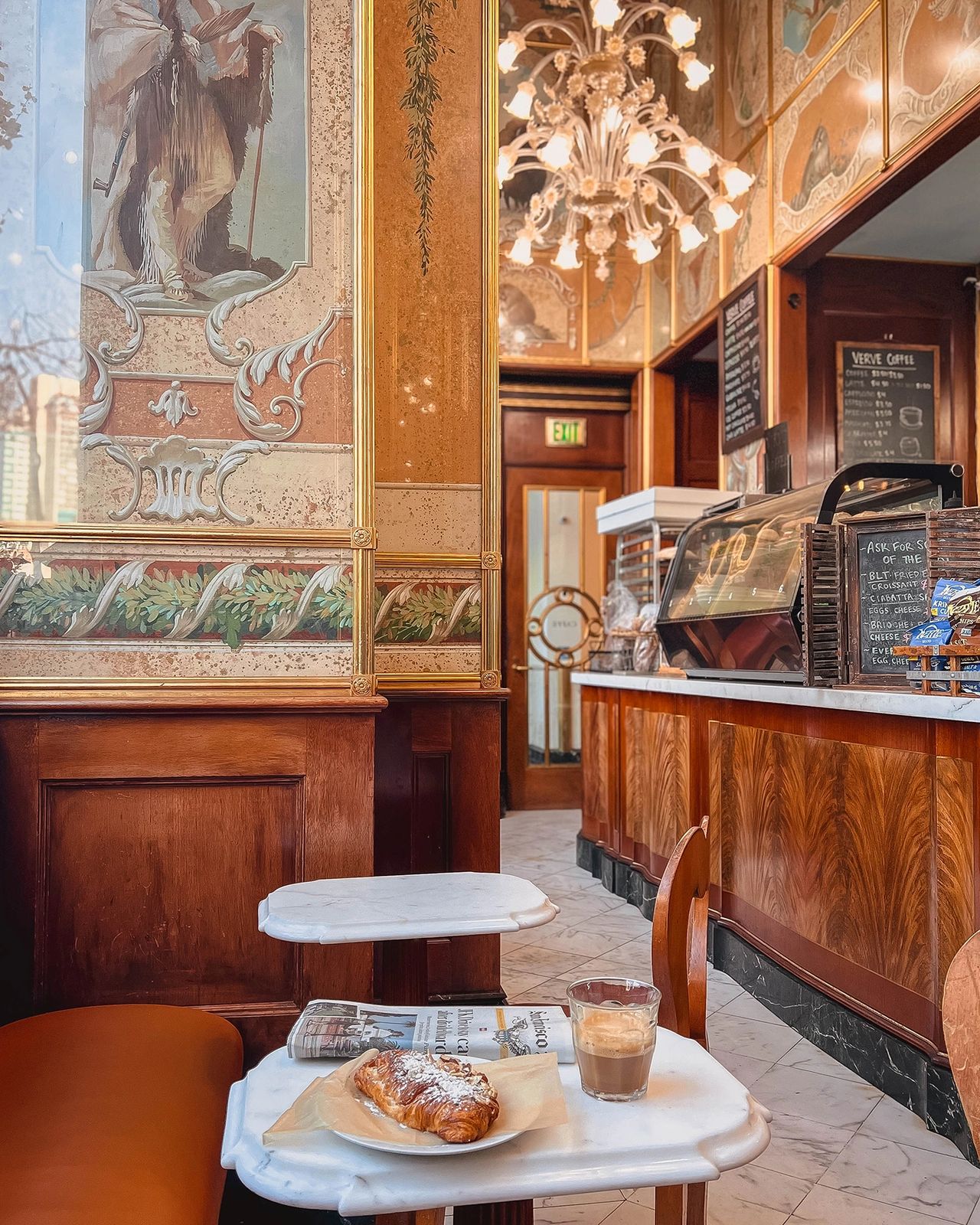 Croissant and coffee on a marble table at La Cuisine Café in San Francisco, surrounded by vintage murals and elegant wood interiors
