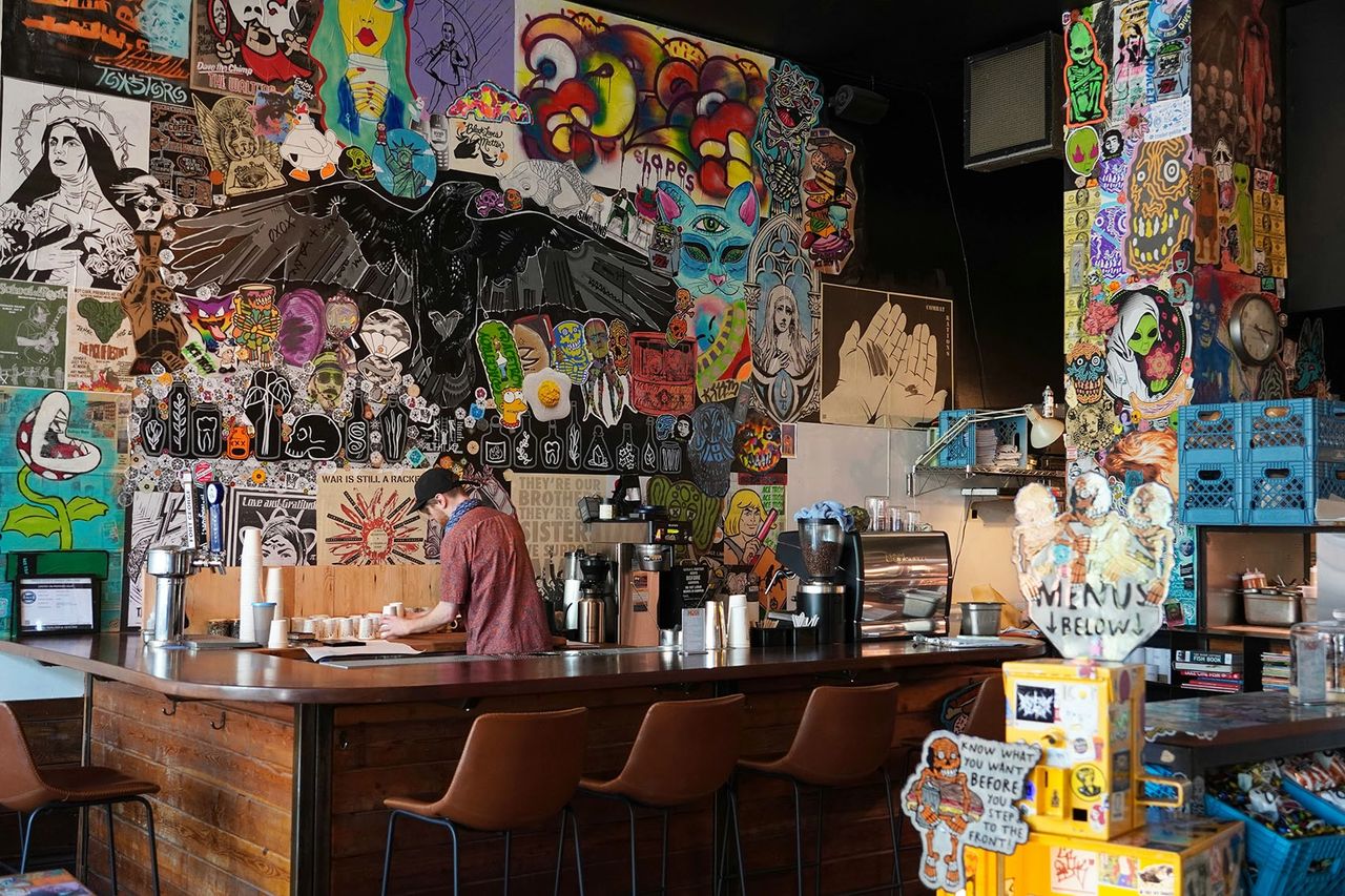 The interior of Lottie & Zula’s café in Portland, Oregon, features a striking collage wall filled with street art, stickers, graffiti-style illustrations, and bold graphic designs. A barista prepares coffee behind a rustic wooden counter, surrounded by vibrant colors and quirky decor. Known for its hearty breakfast sandwiches, excellent coffee, and creative atmosphere, Lottie & Zula’s perfectly embodies Portland’s artistic, laid-back café culture. 