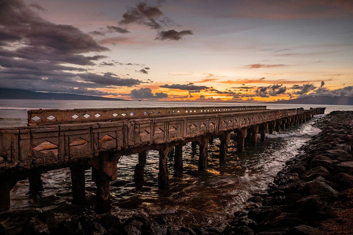 Historic wooden pier stretching into the ocean at golden hour with dramatic clouds over Lahaina, Maui