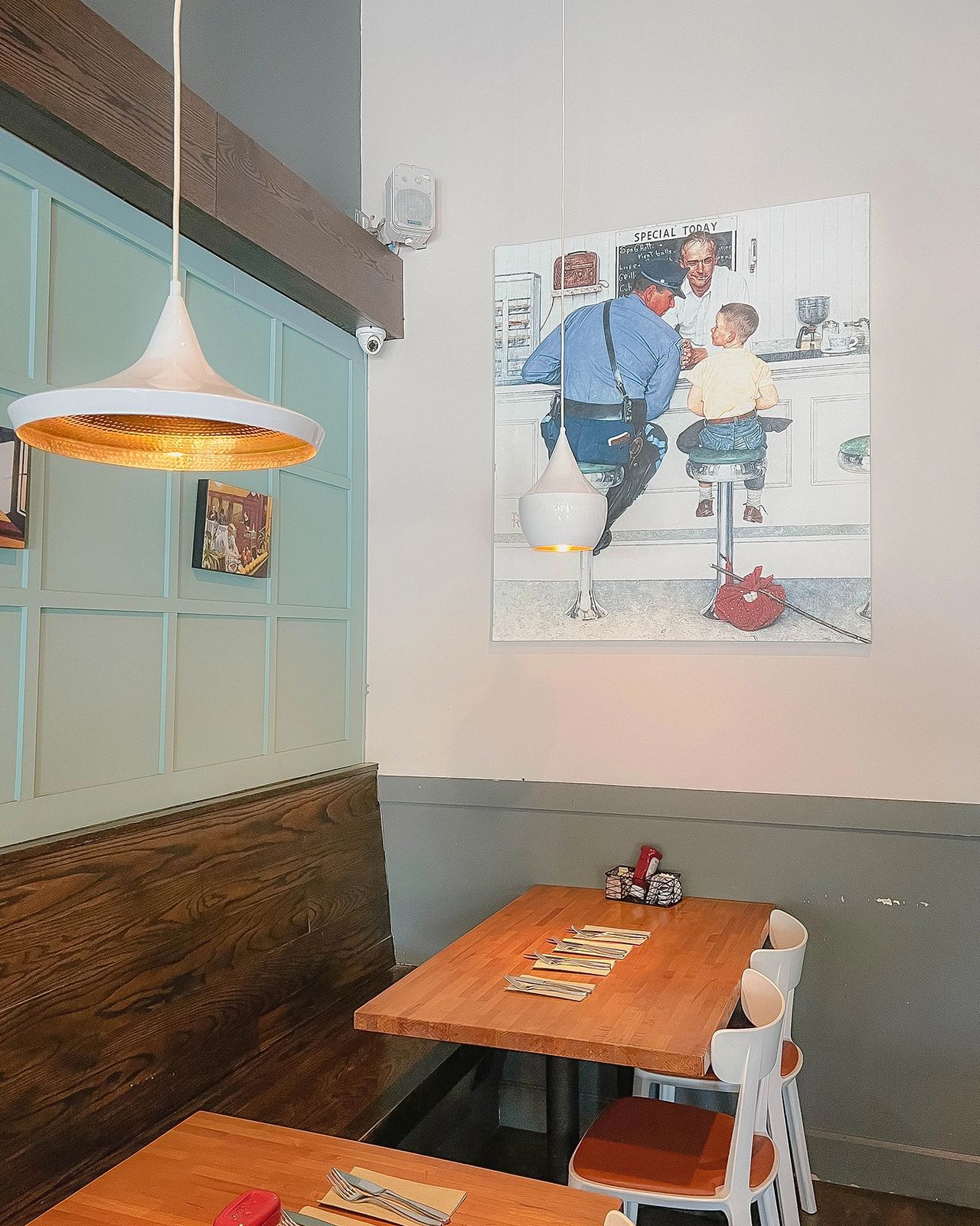 Cozy dining area at Plain Jane in San Francisco, featuring a wooden table setup, retro-style pendant lighting, and vintage Norman Rockwell-style wall art. The warm and inviting design creates the perfect atmosphere for enjoying a relaxed brunch or coffee in one of the city’s favorite neighborhood cafés.