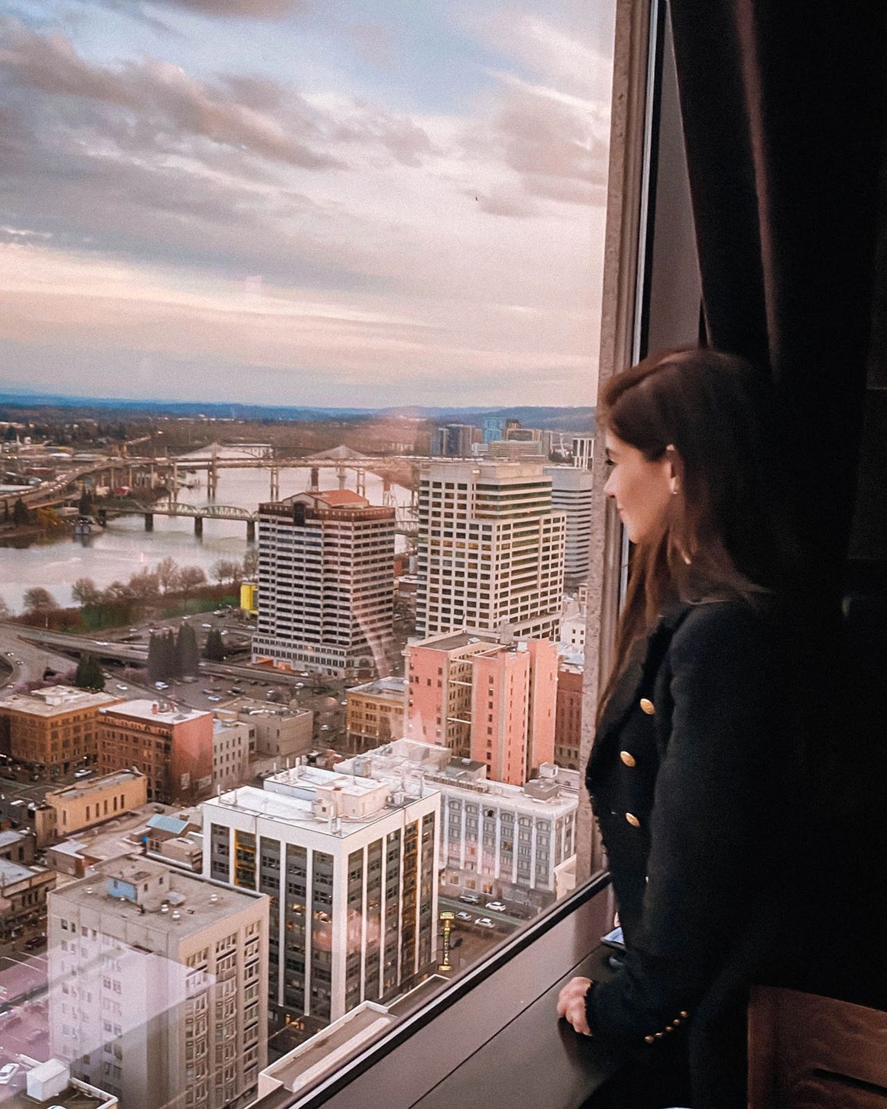 A woman gazes out the window from Portland City Grill on the 30th floor, overlooking downtown Portland, Oregon at sunset. The panoramic cityscape includes the Willamette River, historic bridges, and a mix of modern and historic buildings glowing under soft evening light. Portland City Grill is one of the best places in the city to enjoy sweeping skyline views and sunset drinks. Photo by Nastasia Yakoub