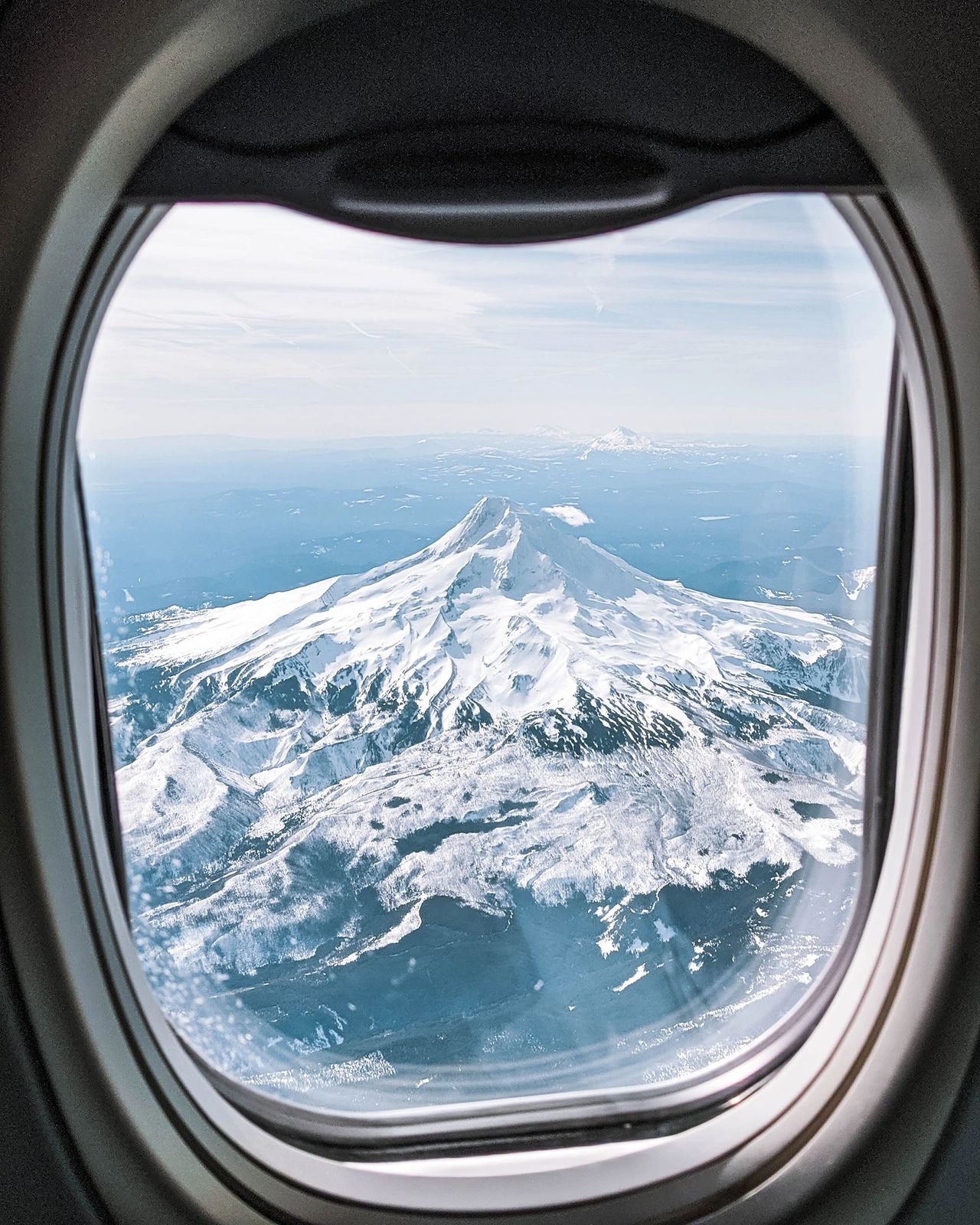  breathtaking aerial view of Mount Hood, Oregon, captured through an airplane window. The snow-covered peak rises majestically above the surrounding landscape under a clear blue sky, offering one of the most iconic sights when flying into or out of Portland. This photo perfectly captures the beauty of the Pacific Northwest and the dramatic scenery that makes Oregon a dream destination for nature and adventure lovers. Photo by Nastasia Yakoub