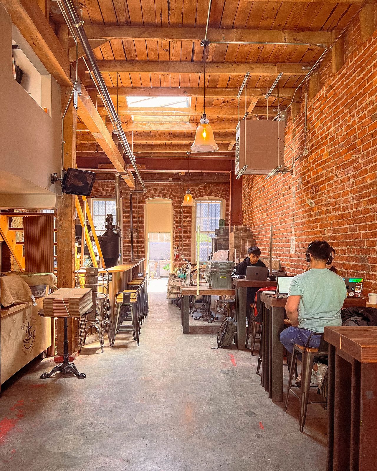 Industrial-style coffee shop interior at Sextant Coffee Roasters in San Francisco with exposed brick walls, wood ceilings, and people working on laptops.